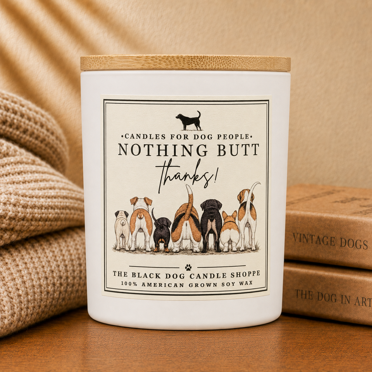 A white candle jar with a bamboo lid sits on a wooden surface, styled with a beige knit blanket on the left and vintage-style dog books on the right. The front label reads 'NOTHING BUT THANKS!' and shows several dog illustrations.