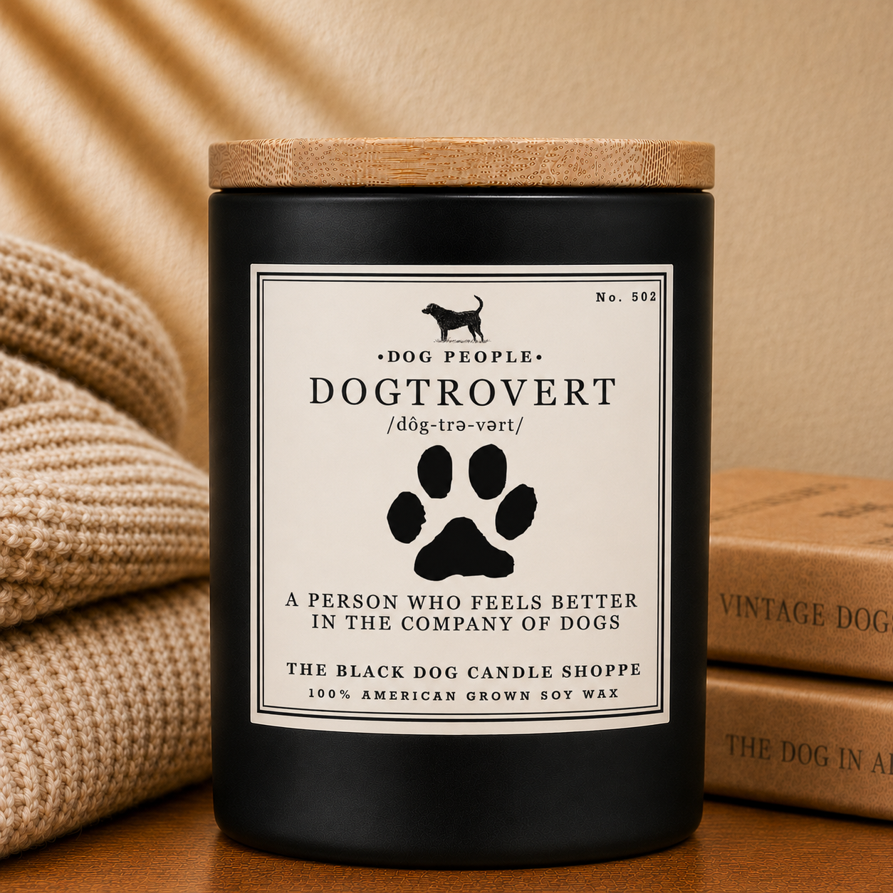 A matte black candle jar with a bamboo lid and a cream label reading 'Dog People Dogtrovert' and 'A person who feels better in the company of dogs,' styled on a wooden surface with a beige knit blanket and vintage books in the background.