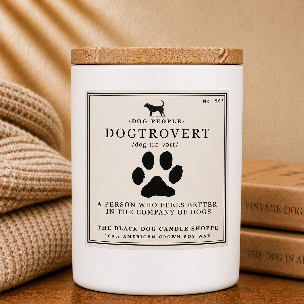 A white soy wax candle jar with a light wood lid and a cream label reading 'Dog People Dogtrovert' with a black paw print, styled on a wooden surface with a beige knit blanket and vintage books in the background.