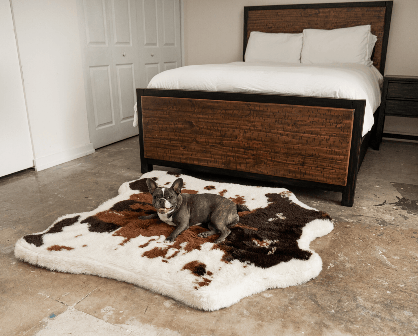 Dog lying on a cowhide rug in front of a wooden bed frame.