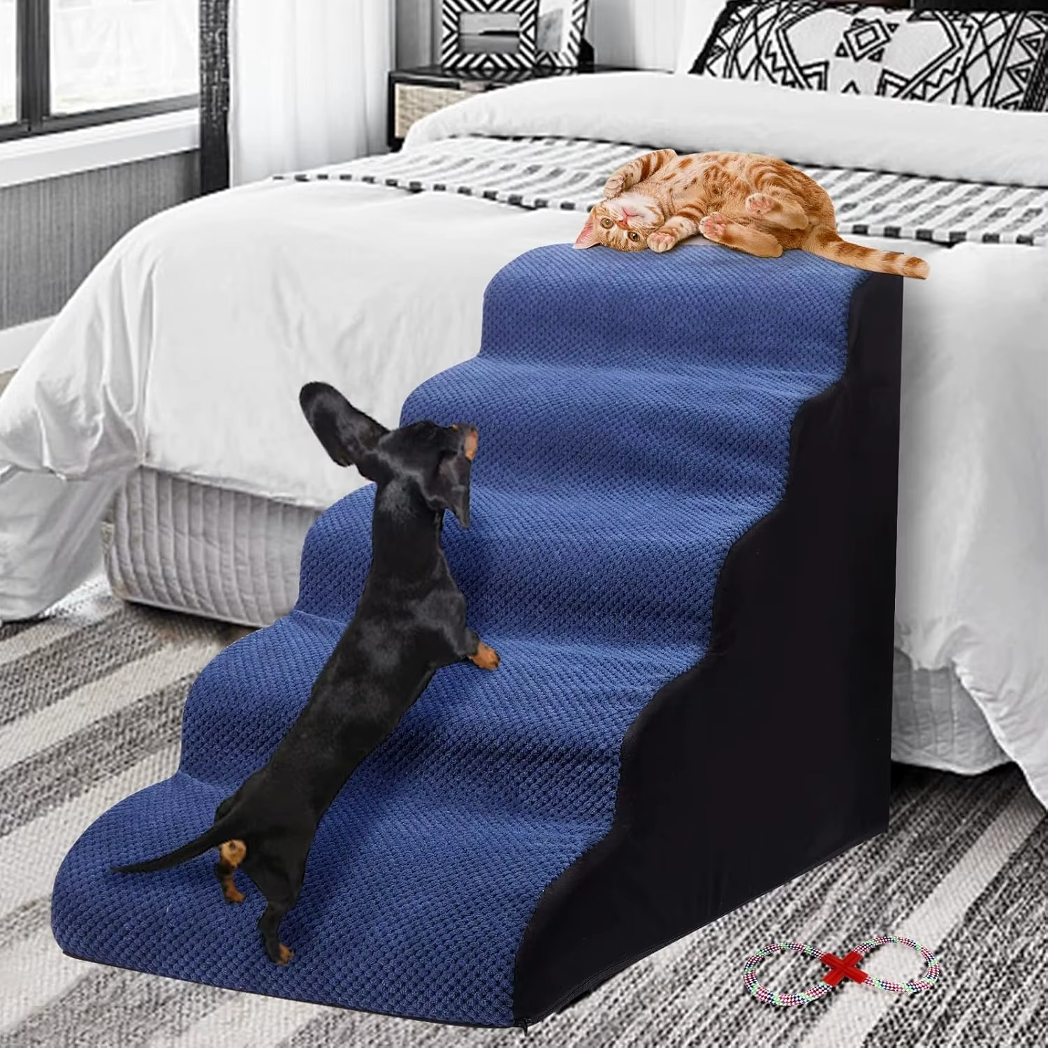 A small white dog sitting on a gray, fabric-covered pet ramp with five steps, placed beside a bed in a home environment.