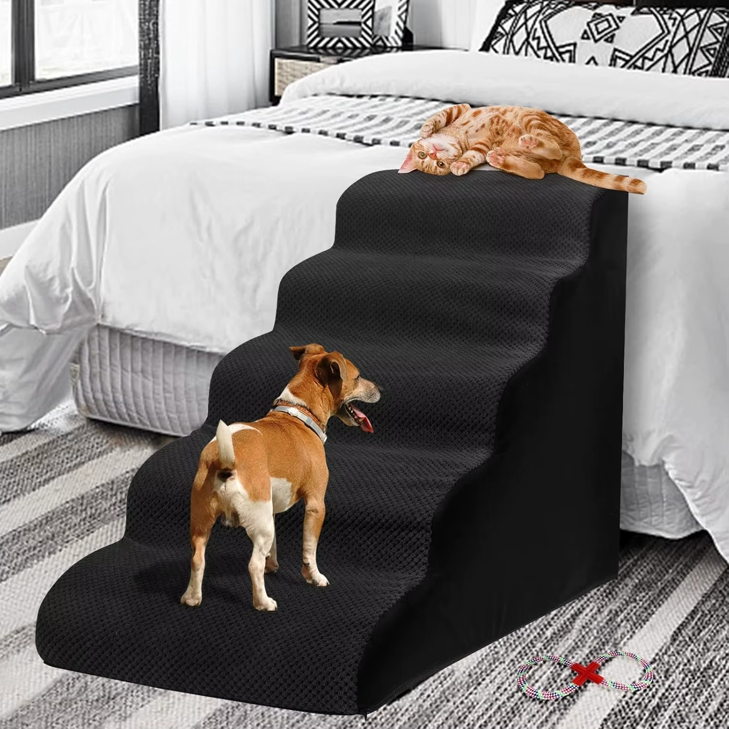 A small white dog sitting on a gray, fabric-covered pet ramp with five steps, placed beside a bed in a home environment.
